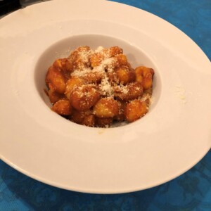 Gnocco dorato con parmigiano in una ciotola bianca su tavolo azzurro. Gnocco dorato in ciotola bianca con spolverata di parmigiano, servito su tavolo azzurro; richiamo alla cucina napoletana dell’Antica Osteria Pisano.