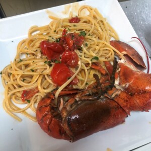 Spaghettoni all'astice con pomodorini e prezzemolo su piatto bianco. Spaghettoni con astice e pomodorini, parmigiano grattugiato, prezzemolo e scorza di limone su piatto bianco; carne d'astice rossa brillante e pasta lunga.