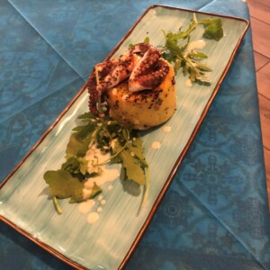 Tortino con polpo e crema su piatto azzurro allungato, decorazione di misticanza. Tortino al centro del piatto decorato con tentacolo di polpo grigliato, salsa cremosa e foglie di misticanza su un piatto azzurro allungato.
