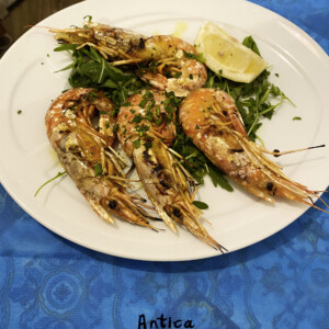Gamberi alla griglia su rucola con limone, Antica Osteria Pisano Gamberi cotti al forno o alla griglia serviti su foglie di rucola con spicchi di limone e prezzemolo, in piatto bianco, contesto di tavolo colorato; rimando alla cucina di Antica Osteria Pisano