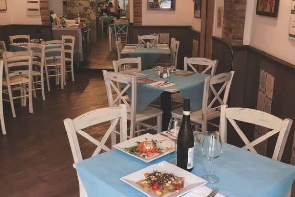 Interni accoglienti di una osteria napoletana con tavoli coperti da tovaglie azzurre, sedie bianche, archi in mattoni e illuminazione calda; presenza di piatti di antipasti e bottiglia di vino